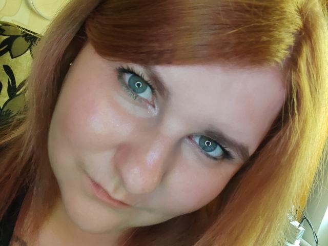 MinistraX has blue eyes, light skin, and reddish-brown hair. She's looking at the camera with a slight smile. Her makeup is subtle, with a bit of eyeliner.