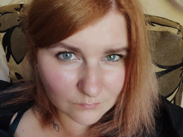 MinistraX has pale skin, red hair, green eyes, and a slight pout with a small nose. Her black top shows a small tattoo near her neck.