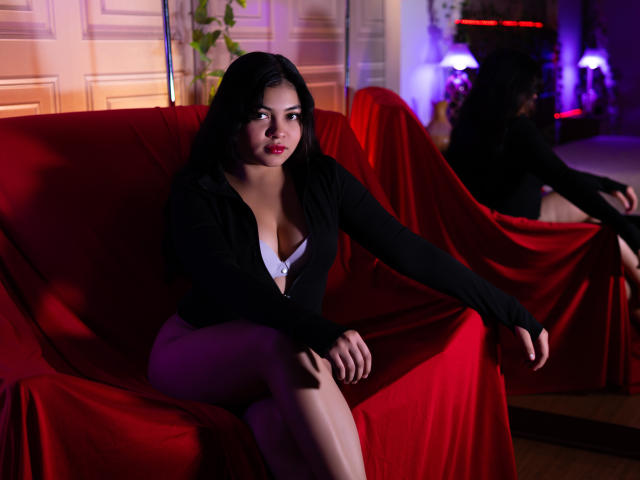 KarinaPerkins with big tits, sitting on a red-covered couch, wearing a black long-sleeve top, white bra, and purple pants, looking at the camera with red lipstick.