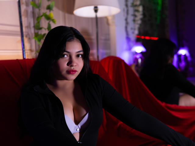 KarinaPerkins with dark hair, red lipstick, and a black zip-up top, showing her tits in a white bra, sitting on a red couch, looking to the side.