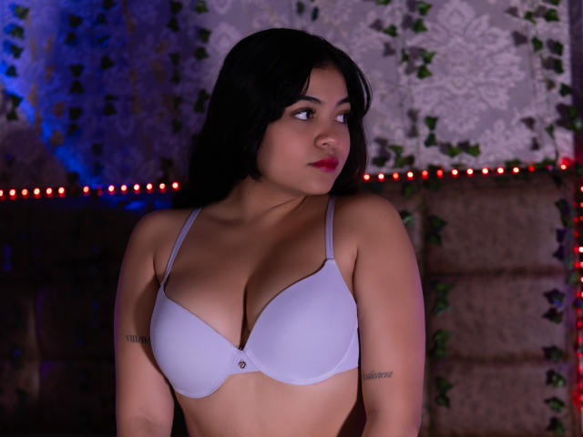 KarinaPerkins with medium brown skin, black hair, red lipstick, white bra, showing her tits, tattoos on arms, looking to the side, red LED strip lights in background.