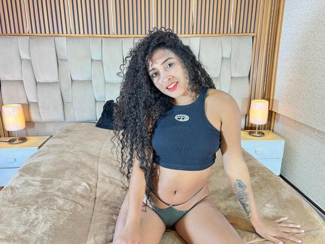 PaulinaRizzo with curly black hair, tan skin, red lipstick, black sports bra, green thong, sitting on a beige bed, tattoo on right forearm, smiling at camera.