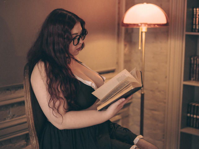 EffyRain with big tits, wearing glasses and a black dress, reads a book under a warm lamp, her long brown hair cascading down. She's sitting, focused, with a book in hand.