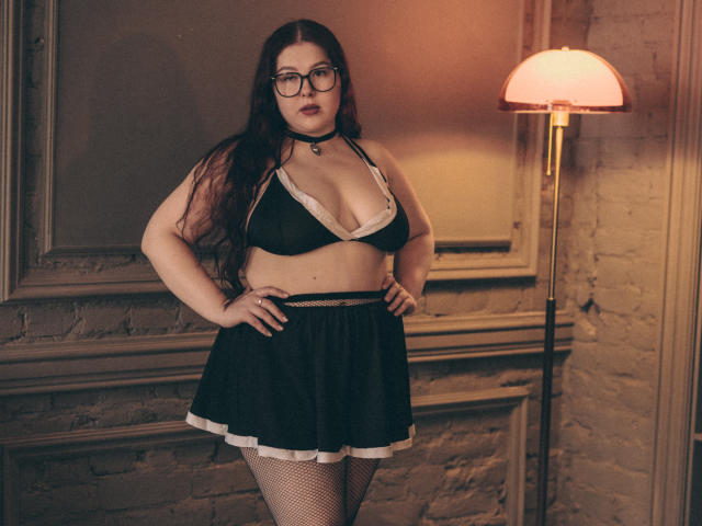 EffyRain with glasses, dark hair, wearing a black bra with white trim, black skirt with white trim, fishnet stockings, black choker, hands on hips, standing in front of a lamp.