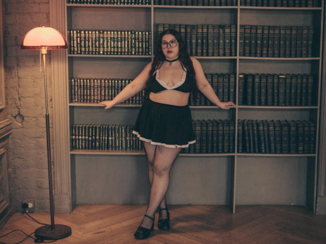 EffyRain with glasses, black and white lingerie, black skirt, white lace trim, black heels, standing against bookshelf, arms out, brick wall, floor lamp, wooden floor.