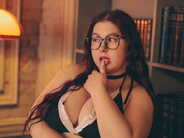 EffyRain with long red hair, glasses, black choker, and black halter top showing big tits, biting finger, looking seductive, warm lighting, bookshelf in background.