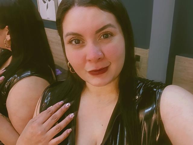 RachelCarlton with black hair, wearing a shiny black leather top, showing her tits in a mirror selfie, smiling, pink nail polish, gold hoop earrings, looking at the camera.