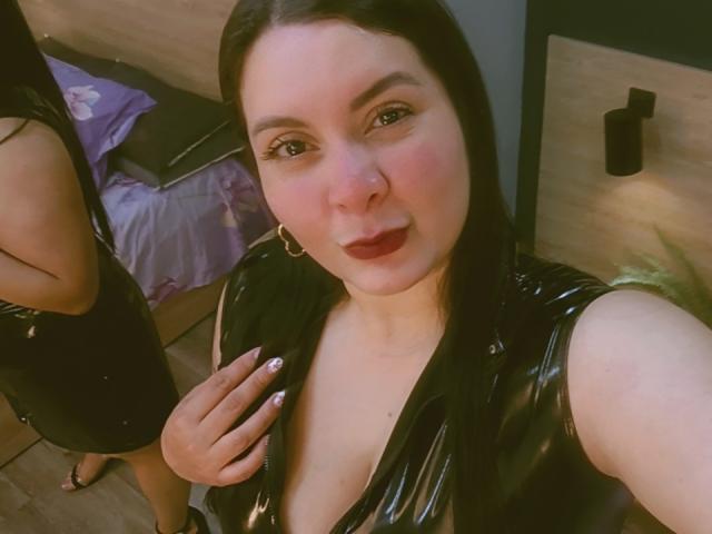 RachelCarlton with black latex dress, medium tits, pink blush, red lipstick, black hair, gold hoop earrings, holding dress, standing next to another woman in black dress.