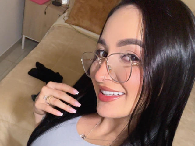 PamelaLiss with long black hair, glasses, nose ring, pink lipstick, smiling, grey top, ring on right hand, black sleeve, beige bed, light skin, pink nail polish, smiling.