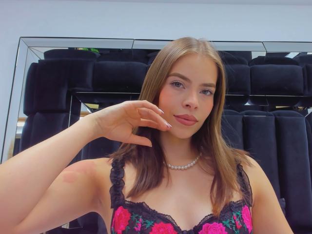 AnnaFer mit glattem, braunem Haar, rosa Lippenstift und einem schwarzen Spitzenkleid mit Blumenmuster, die ihre Brüste zur Schau stellt, wobei sie eine Hand in die Nähe ihres Gesichts bringt, und eine Perlenkette trägt..