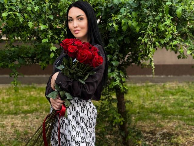 Xioni with long black hair, holding a bouquet of red roses, wearing a brown shirt and white patterned skirt, standing in front of a green tree.