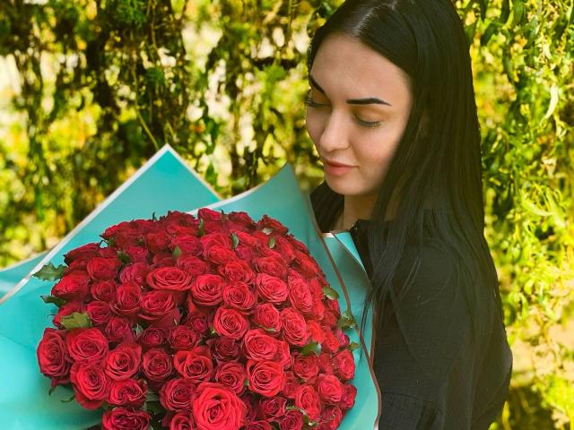 Xioni with long black hair, holding a large bouquet of red roses, wrapped in blue paper, has a soft smile and looks down. She's wearing a black shirt.
