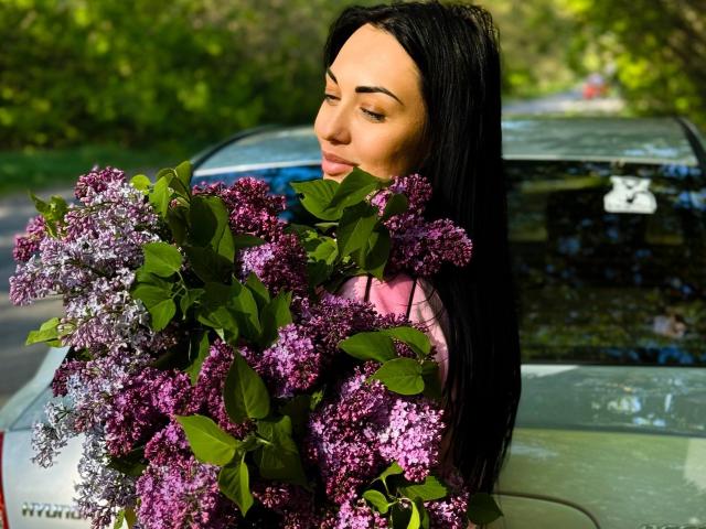 Xioni with long black hair holding a bunch of purple lilacs, standing in front of a green car, smiling gently. Her tits are partially visible under the flowers.