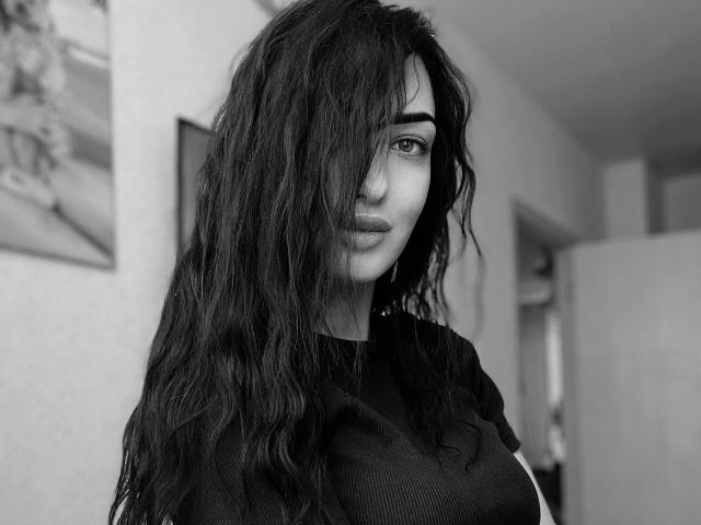 Black-and-white photo of Xioni with long, wavy, dark hair covering one eye, wearing a black shirt, showing a subtle, confident smile, with visible breasts in the foreground.