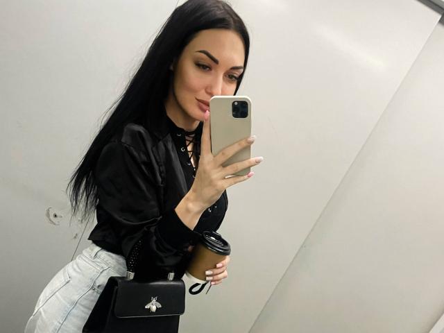 Xioni with long black hair, black lace-up top, white high-waisted jeans, holding coffee cup and black bag, taking selfie in mirror with iPhone, pale skin, pink nails.