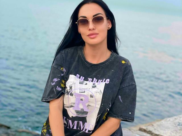 Xioni has long black hair, wears sunglasses, and a dirty black t-shirt with a graphic print. Her ass is not visible. She's standing by the water.