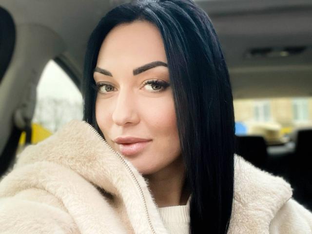 Xioni has black hair, brown eyes, and is wearing a fluffy beige coat. She's looking at the camera with a slight smile, showing a bit of her pink lips.