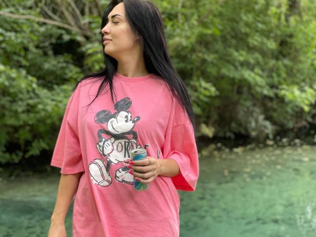 Xioni with long black hair, pink t-shirt featuring Mickey Mouse, holding water bottle, standing by green water, looking to the side, sexy, cute, casual, outdoors.