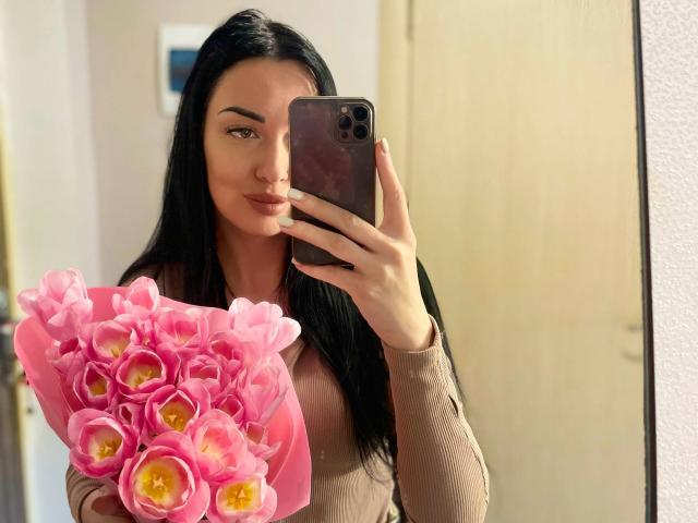 Xioni with long black hair, holding a pink bouquet of tulips, taking a mirror selfie with a brown phone, wearing a beige long-sleeve top, looking slightly smug.