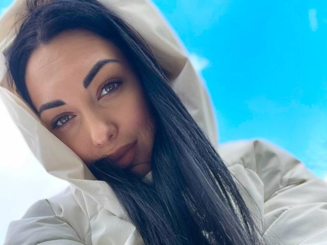 Xioni has dark, straight hair, brown eyes, and thick eyebrows. She's wearing a white hooded jacket, looking down with a slight smile. Her tits are not visible.