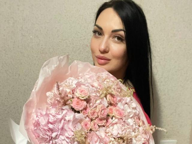 Xioni with long black hair, holding a pink rose bouquet, has a subtle smile, and a cute nose. She's wearing a pink shirt, and looking at the camera.