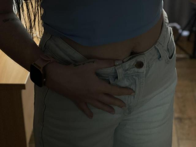 Xioni's hand is gripping her white jeans, pulling them down slightly to reveal her soft, pale belly. She wears a blue top and a pink smartwatch.