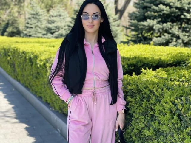 Xioni with long black hair, wearing pink tracksuit, black vest, and glasses, standing by green bushes, hands in pockets, holding black bag, sunny day.