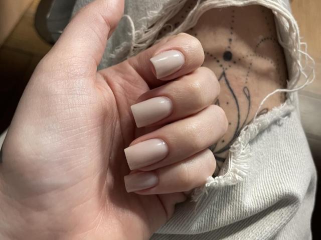 Xioni's hand with long, nude-painted nails, showing a tattoo of a constellation on their chest, through a ripped gray shirt. Their fingers are slightly curled.