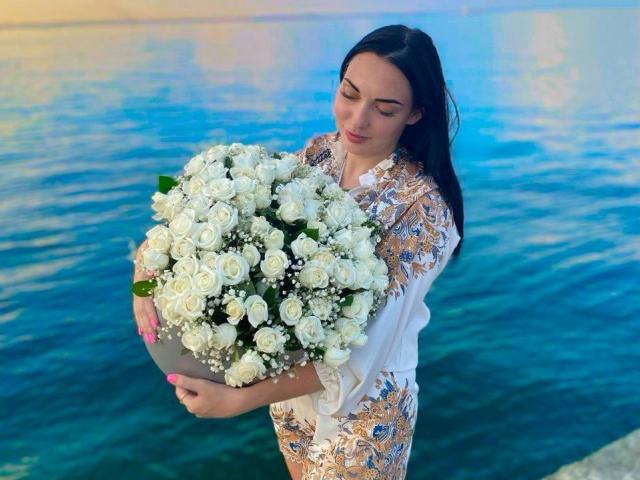 Xioni with dark hair, holding a large bouquet of white roses and baby's breath, wearing a white and gold floral dress, standing by a blue water body.