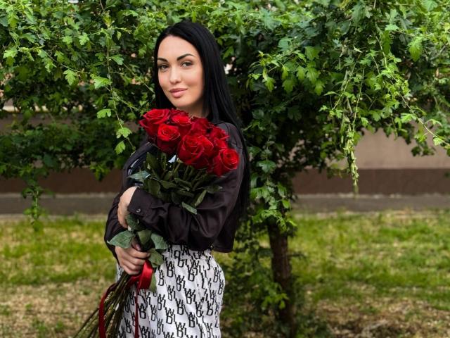 Xioni with black hair, holding a bouquet of red roses, wearing a black shirt and white patterned skirt, standing under a green tree. Her tits are small, she's cute.