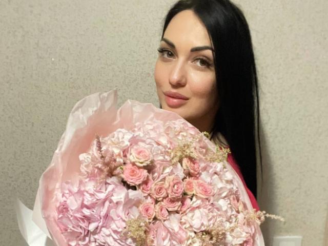 Xioni with long black hair, holding a pink bouquet of roses, has a slight smile, and is looking at the camera with a subtle, sexy expression.