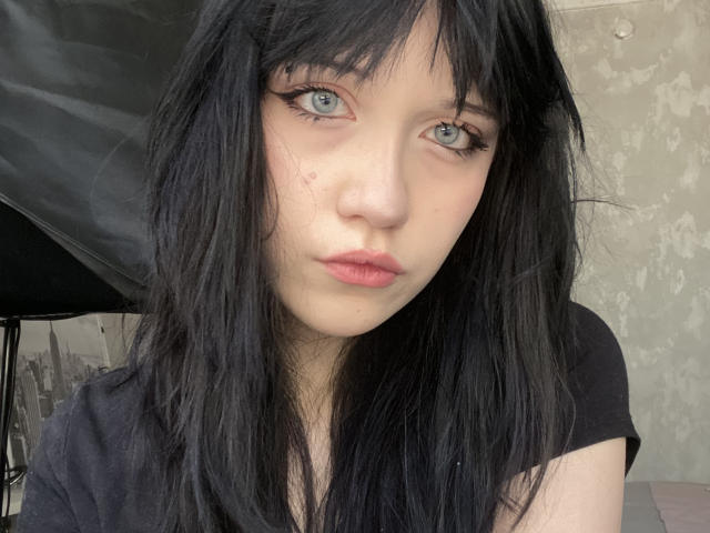 SantinaSahm has pale skin, black messy hair, blue eyes, and pink lips, with a small zit on her cheek. She's wearing a black shirt, looking serious.