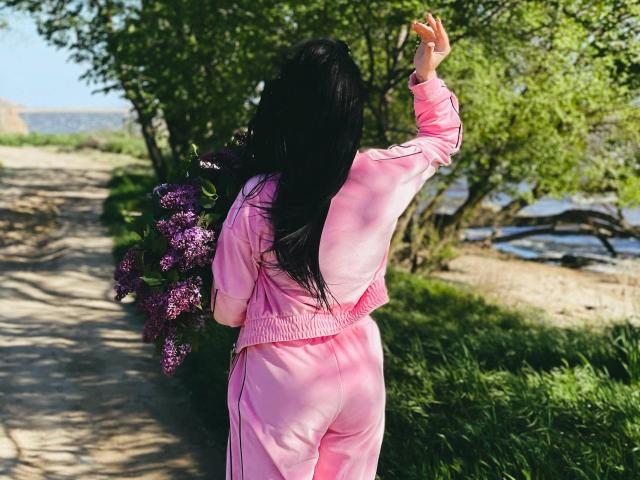 Xioni with long black hair, wearing pink tracksuit, holding purple flowers, raising arm, standing on dirt path, green bushes, and trees in background.