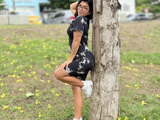 ValeryPierce leans against a tree, wearing a black and white dress, white sneakers, glasses, and tattoos on her arm. Her ass is prominent, looking back with a smile.