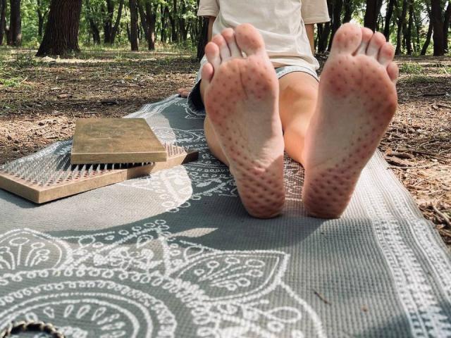 TenerliRize's bare feet with pink soles are in the foreground, soles facing camera, sitting on a patterned mat with a wooden comb and book to the left.