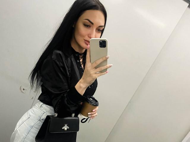 Xioni with long black hair, black blouse, and white high-waisted pants, holding phone and coffee cup, tongue out, black cross-body bag, in mirror selfie.
