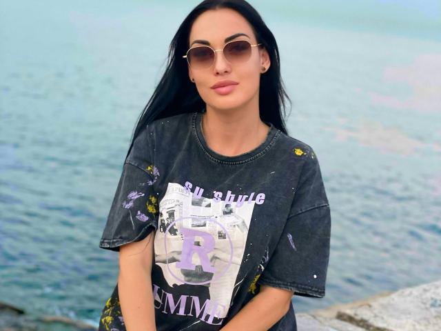 Xioni with long black hair, wearing a dirty black R&L t-shirt with a purple "R" and yellow flowers, and round sunglasses, standing by a blue water body.