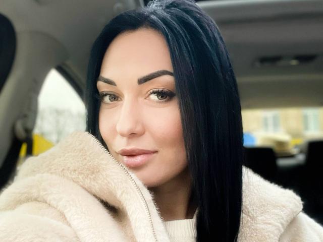 Xioni has black hair, brown eyebrows, and wears a fluffy beige coat in a car. She has a slight smile and subtle makeup. Her tits are not visible.