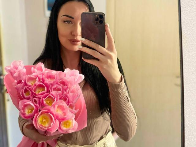 Xioni with long black hair, holding a pink bouquet of tulips, in a beige long-sleeve top, taking a mirror selfie with an iPhone. Boobs, ass, tits, pussy, not visible.