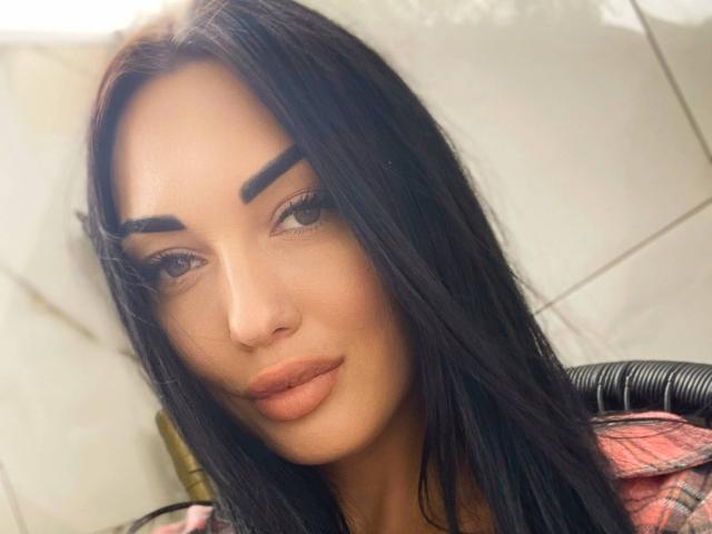 Xioni has long black hair, thick eyebrows, and full, slightly pouty lips with a subtle smile. She's wearing a pink plaid shirt. Her skin is tan.