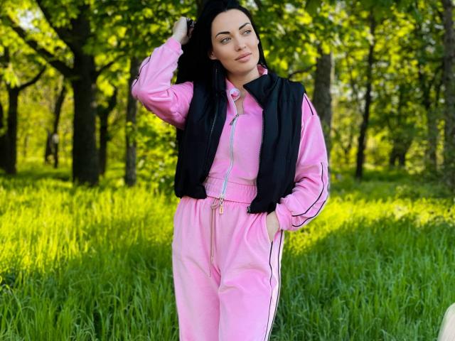 Xioni with black vest over pink tracksuit, hand in pocket, green grass, and trees, black hair, slight smile, sunlight, relaxed pose. She's cute, with a sexy, slim build.