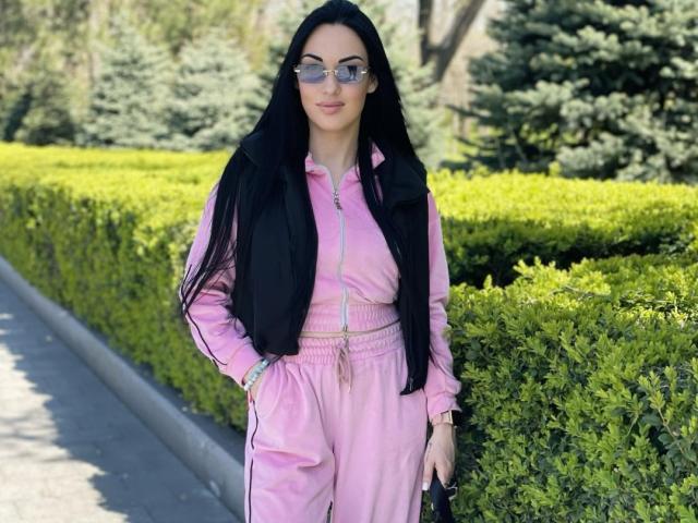 Xioni with long black hair, wearing pink track suit, black vest, and glasses, hands in pockets, standing on sidewalk with green hedge. She has a cute, sexy look.