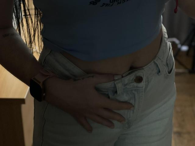 Xioni's hand is pulling down her white jeans, exposing her soft, pale belly and slightly visible pussy. She wears a blue top and a pink smartwatch.