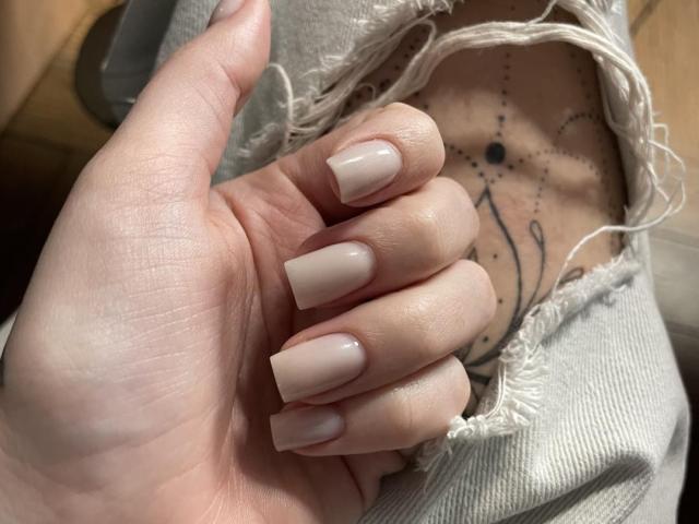 Xioni's hand with pale pink, almond-shaped nails, showing a tattoo on their chest with a black star and line pattern, under a frayed gray sleeve.