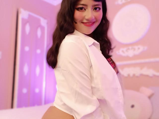 NekoPam with wavy dark hair, wearing a white shirt, plaid necktie, smiling, showing her ass, pink room, soft pink and white decor, plush toy in background.