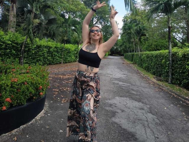 AlinaMatei stands on a wet, tree-lined path, arms raised, flashing rock signs, with tits visible, wearing a black sports bra and patterned skirt, tattoos on chest and lower belly.