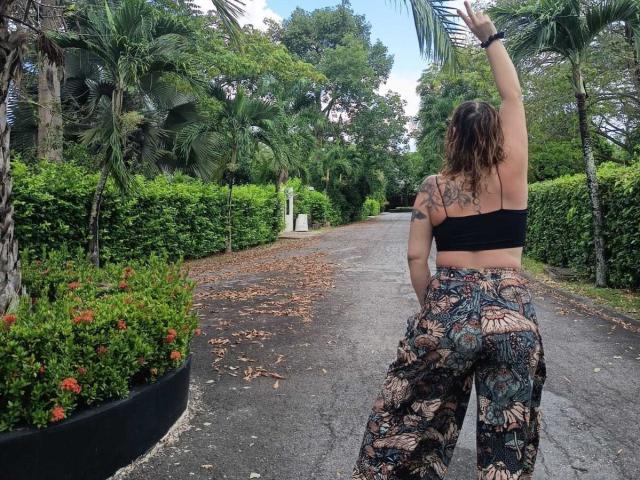 AlinaMatei with tattoos on her back, black bra, and floral pants, raising her arm, standing on a gravel path, palm trees and greenery in the background.