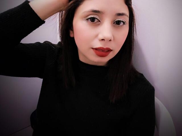 Stephanie Sun, con cabello castaño oscuro, lápiz labial rojo, suéter negro, mano en el cabello, sentada, piel clara, labios rojos ligeramente entreabiertos, nariz pequeña, maquillaje sutil, mirando a la cámara, linda..