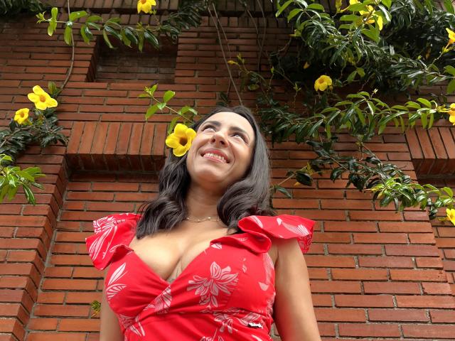 JadeFoster with medium tits, wearing a red floral dress, smiling up at yellow flowers, dark wavy hair, and a necklace, standing in front of a red brick wall.