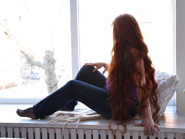 OVanessaHotly with long red hair, wearing a purple top and blue jeans, sitting on a windowsill, legs crossed, back to the camera, butt up, hands on knee and window.